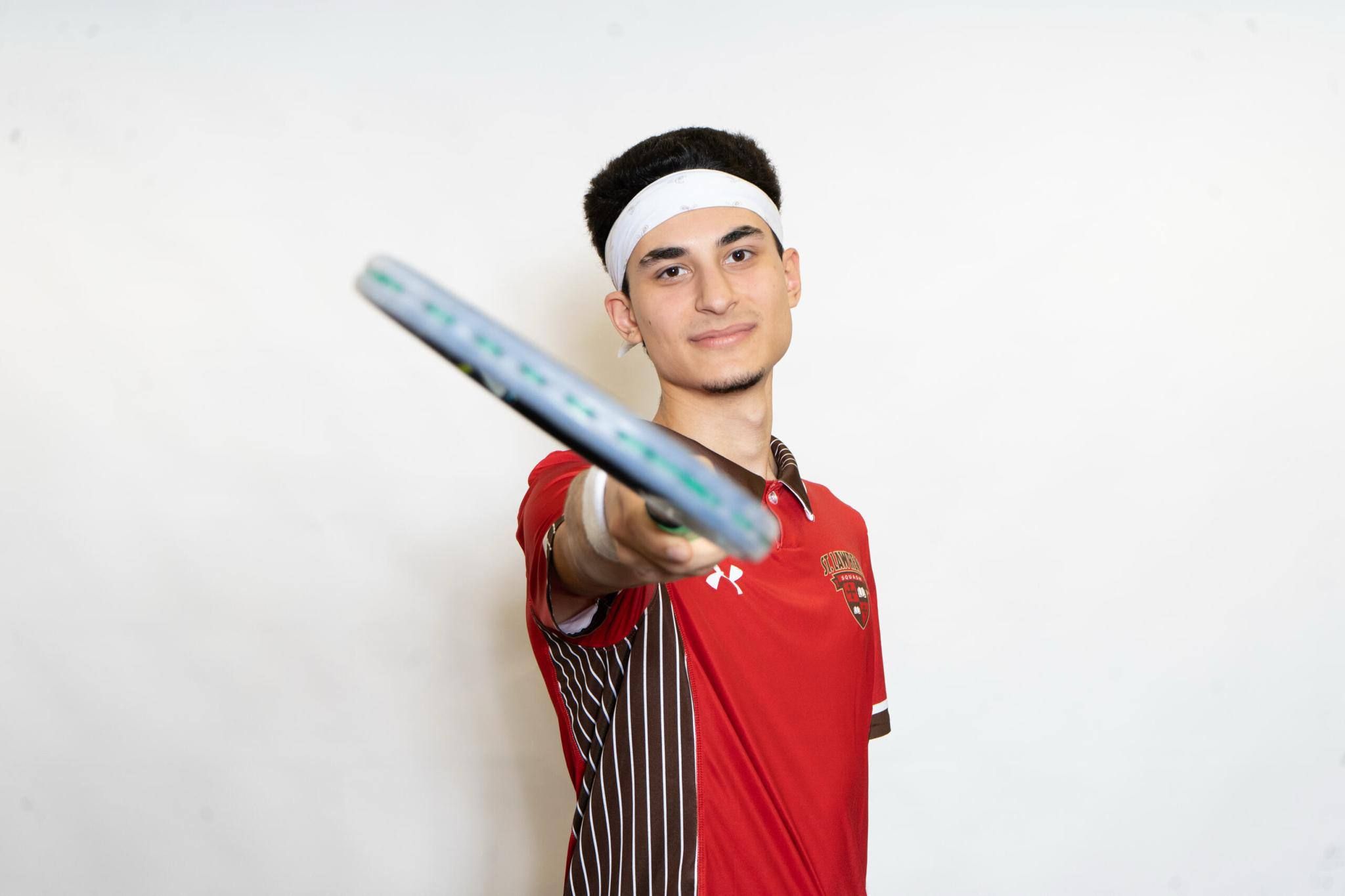 Emirhan Sen in a red St. Lawrence squash shirt and white headband, holding his racket out toward the camera